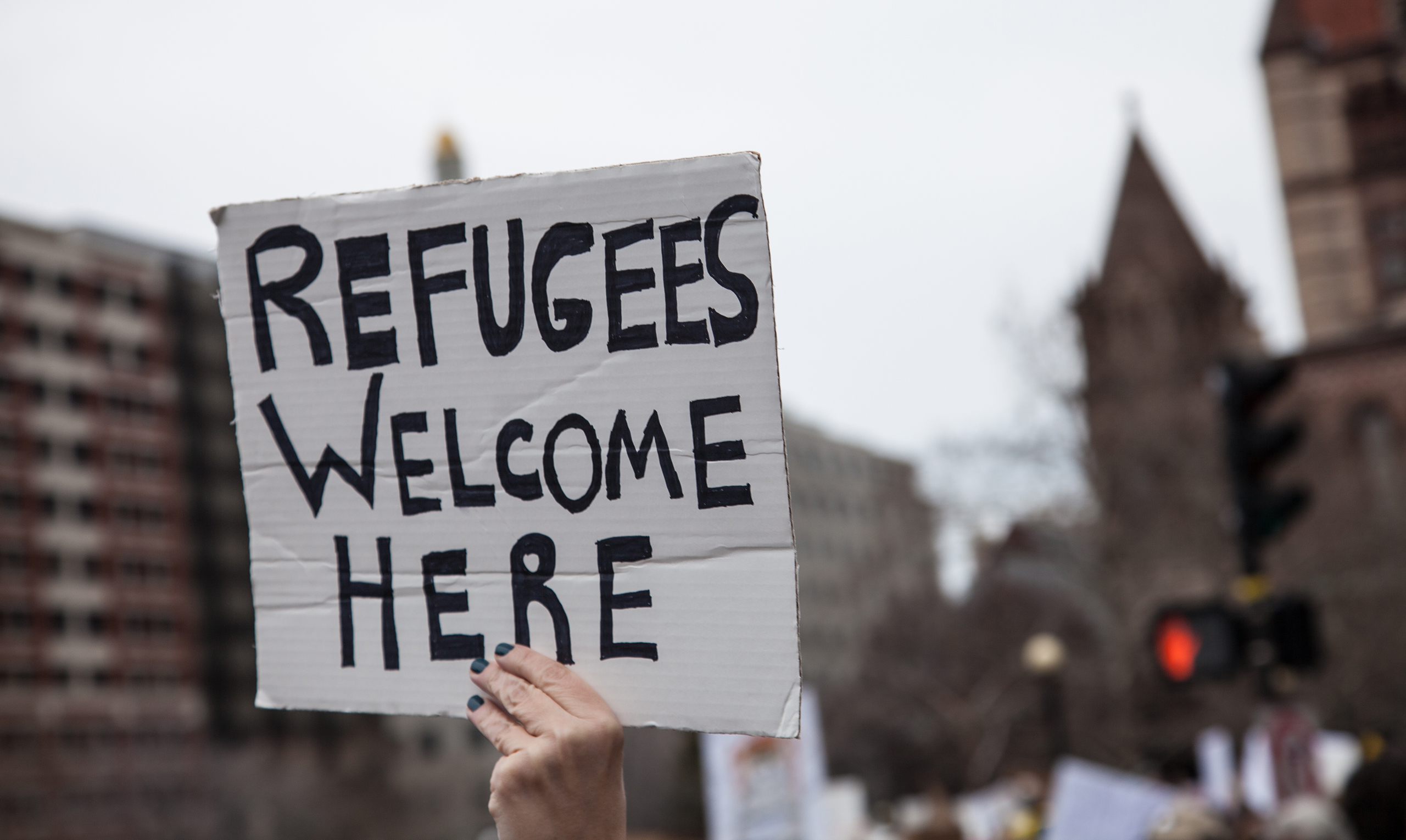 A sign is held above a crowd of protesters that reads "Refugees Welcome Here"