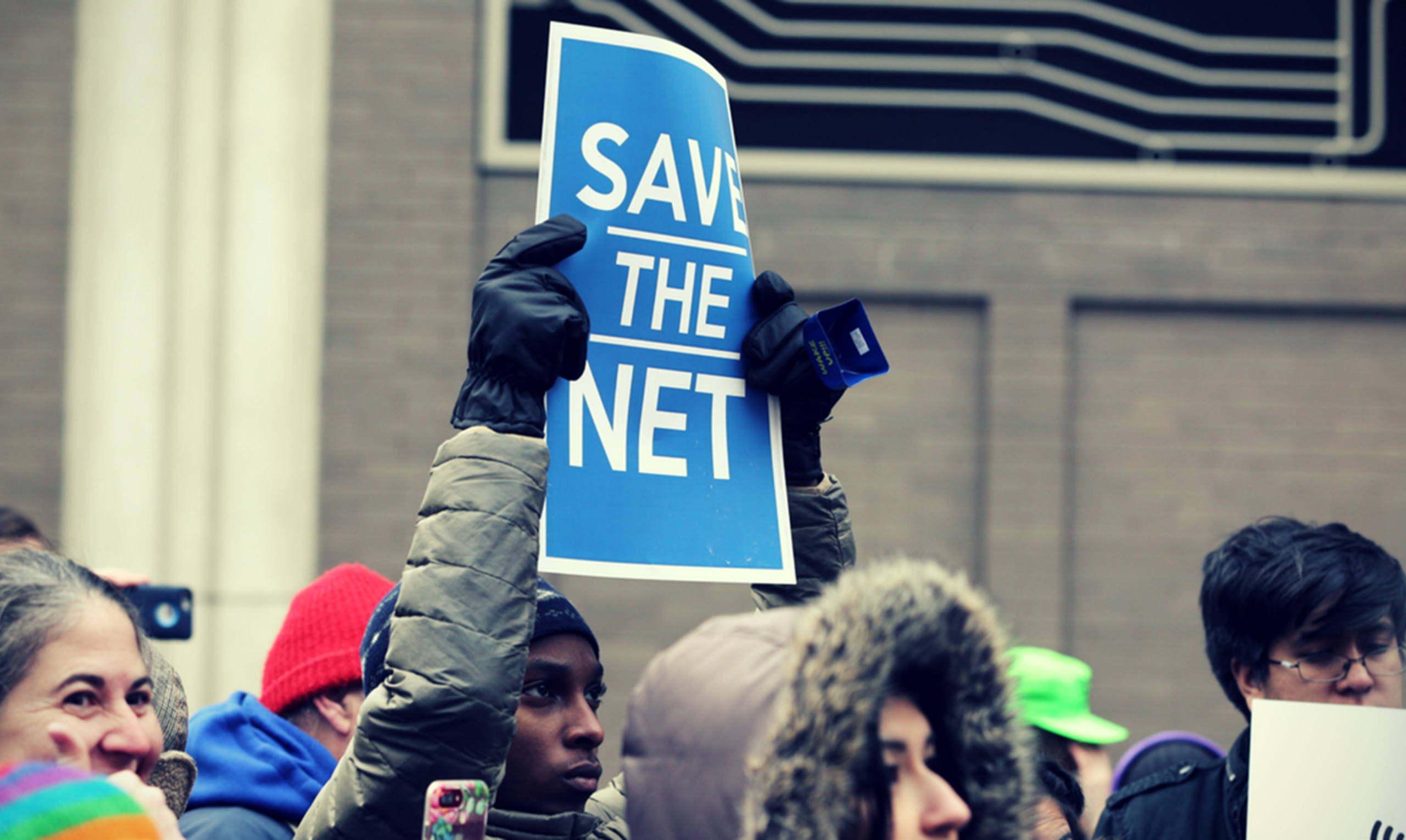 A person at a protest holds a sign reading "Save the Net"